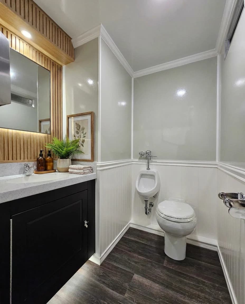 Men's suite: black shaker vanity with quartz counter, backlit wood-slat mirror, fern and amber soap bottles, porcelain toilet and wall-hung urinal